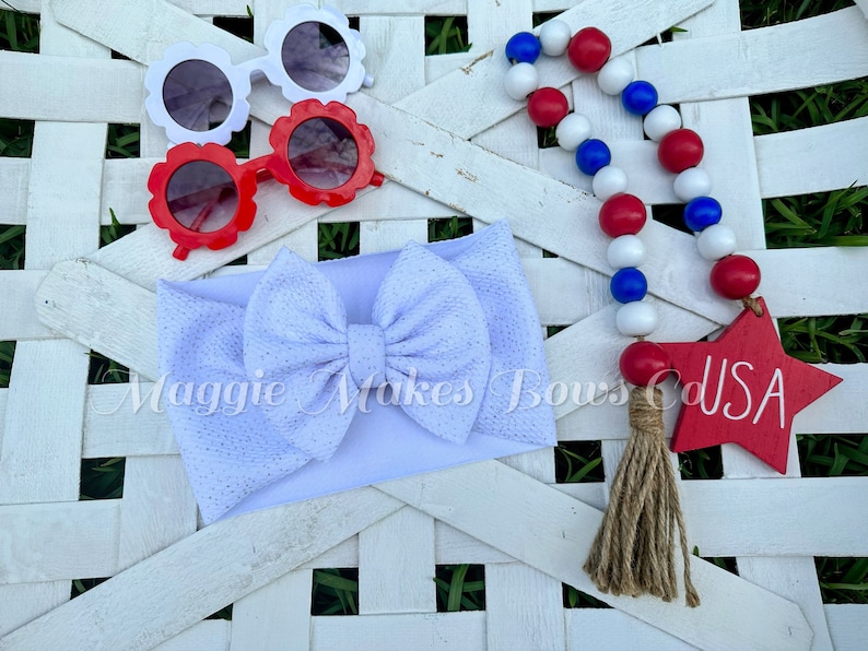 May include: White headband with a white bow, red and white flower-shaped sunglasses, and a red, white, and blue beaded garland with a wooden star that says "USA".