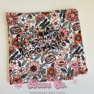 May include: A white fabric with a pink and orange floral print. The fabric is folded in half and a matching bow is placed on top. The fabric has the text "Bows Co. Handmade Boutique" at the bottom.