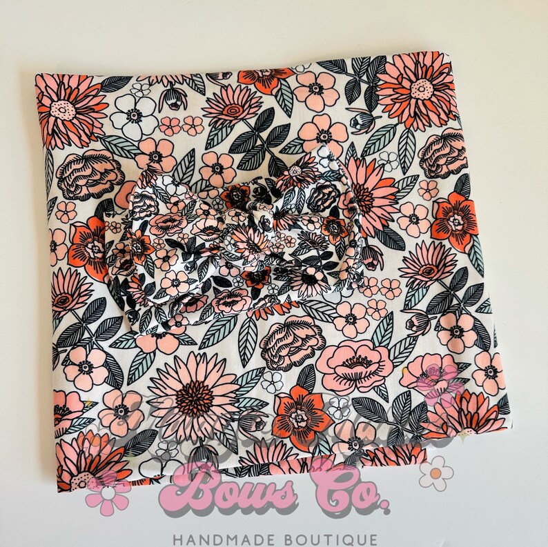 May include: A white fabric with a pink and orange floral print. The fabric is folded in half and a matching bow is placed on top. The fabric has the text "Bows Co. Handmade Boutique" at the bottom.