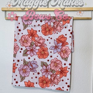 May include: A white fabric with a floral print featuring pink, orange, and purple flowers with green leaves and white stars. The fabric is hanging on a wooden hanger.