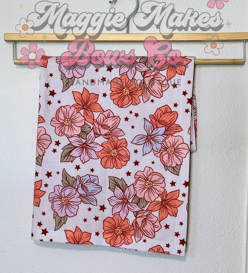 May include: A white fabric with a floral print featuring pink, orange, and purple flowers with green leaves and white stars. The fabric is hanging on a wooden hanger.