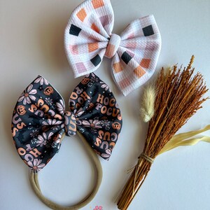May include: Two decorative hair bows. One bow features a black background with orange and pink floral and text that says "Hocus Pocus". The other bow has a white background with a checkered pattern of orange, pink, and black squares. A bundle of dried wheat is also present.