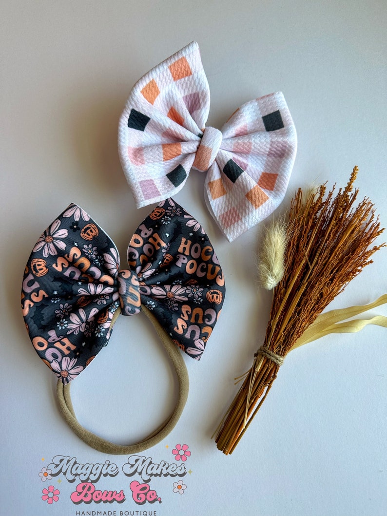 May include: Two decorative hair bows. One bow features a black background with orange and pink floral and text that says "Hocus Pocus". The other bow has a white background with a checkered pattern of orange, pink, and black squares. A bundle of dried wheat is also present.