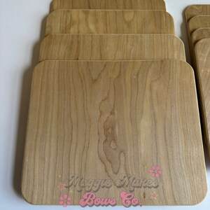 May include: A stack of light-colored wooden boards with rounded corners. The wood grain is visible, and the boards are arranged in a neat, overlapping pattern. The text "Maggie Makes Bows Co. Handmade Boutique" is printed on the boards.