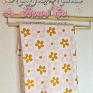 May include: A pink and white fabric with a checkered pattern and orange daisy flowers. The fabric is hanging on a wooden hanger with the text "Maggie Makes Bows Co. Handmade Boutique".