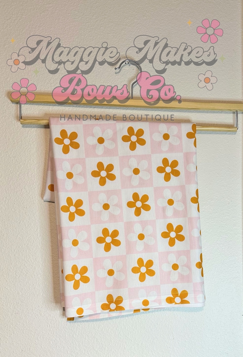 May include: A pink and white fabric with a checkered pattern and orange daisy flowers. The fabric is hanging on a wooden hanger with the text "Maggie Makes Bows Co. Handmade Boutique".