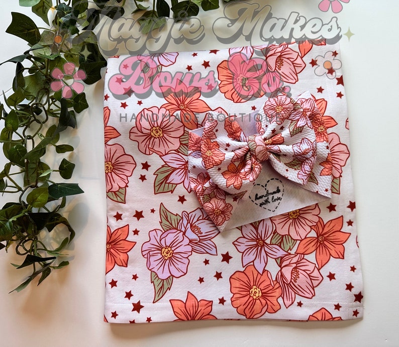 May include: A white fabric baby blanket with a floral print in shades of pink, orange, and green. The blanket has small red stars scattered throughout the print. A pink and orange floral bow is on top of the blanket. The bow has a small white tag that says "Handmade with love".