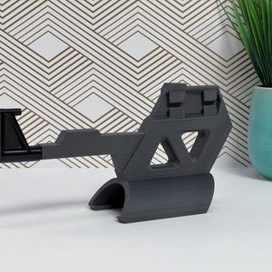 May include: A dark gray, geometric-shaped stand with a black hook. The stand has a curved base and a triangular cutout. A black hook extends from the stand. The stand is on a white surface.