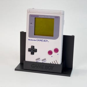 May include: A white Nintendo Game Boy handheld console in a black stand. The Game Boy features a grey screen, a black directional pad, pink buttons, and the text "GAME BOY" printed on the front. The stand is black and has the text "GAME BOY" on the base.