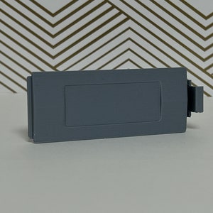 May include: A gray plastic battery cover with a rectangular opening.