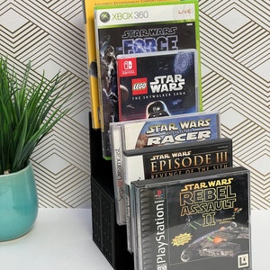 May include: A black video game storage stand with a variety of Star Wars video games. The games include Star Wars: The Force Unleashed, LEGO Star Wars: The Skywalker Saga, Star Wars: Episode I Racer, Star Wars: Episode III - Revenge of the Sith, and Star Wars: Rebel Assault II - The Hidden Empire.