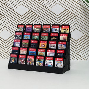 Puede incluir: Un soporte de plástico negro contiene una colección de cartuchos de juegos de Nintendo Switch. Los cartuchos están dispuestos en filas y columnas, con los títulos de los juegos visibles. El soporte está sobre una superficie blanca.