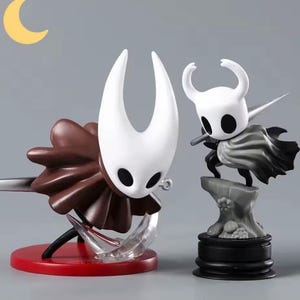 May include: Two detailed figurines of characters from the video game Hollow Knight. One figure has a large white head with horns and a brown cape. The other is a smaller white figure with a cape, standing on a rock base. The figures are on a gray background.
