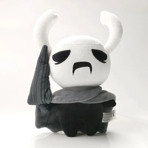 Può includere: Un giocattolo di peluche bianco con occhi neri e una smorfia. Il giocattolo indossa un mantello grigio e ha una spada nera. Il giocattolo è un personaggio del videogioco Hollow Knight.