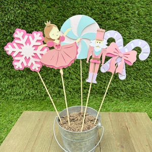 May include: A collection of decorative paper cutouts on wooden sticks, including a pink snowflake, a ballerina, a blue and white swirl, a nutcracker, and a candy cane with a bow. The cutouts are arranged in a small metal bucket filled with sand, set against a green background.