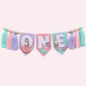 May include: A festive banner with the word "ONE" spelled out on colorful pennants. Each pennant features a different design, including a ballerina, a lollipop, and a toy soldier. The banner is adorned with tassels in shades of teal, pink, and lavender.