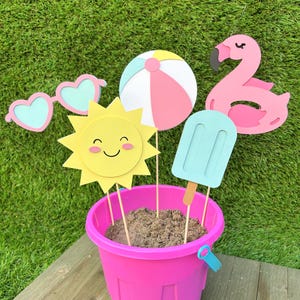 May include: A pink bucket filled with sand holds several summer-themed cutouts. These include heart-shaped sunglasses, a beach ball, a smiling sun, a popsicle, and a flamingo. The cutouts are in pastel colours and are on wooden sticks.
