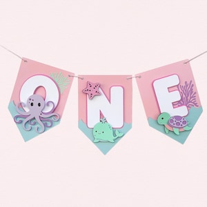 Pu&ograve; includere: Una ghirlanda decorativa con la scritta "ONE", ogni lettera su un gagliardetto rosa con base blu. La "O" presenta un polpo viola, la "N" una balena verde e una stella marina, e la "E" una tartaruga.
