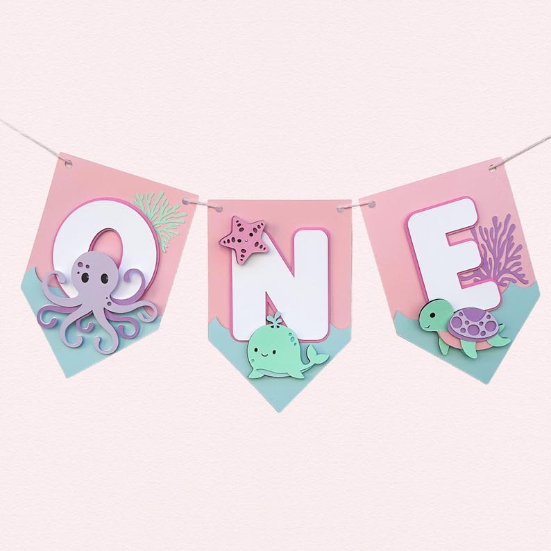 Pu&ograve; includere: Una ghirlanda decorativa con la scritta "ONE", ogni lettera su un gagliardetto rosa con base blu. La "O" presenta un polpo viola, la "N" una balena verde e una stella marina, e la "E" una tartaruga.
