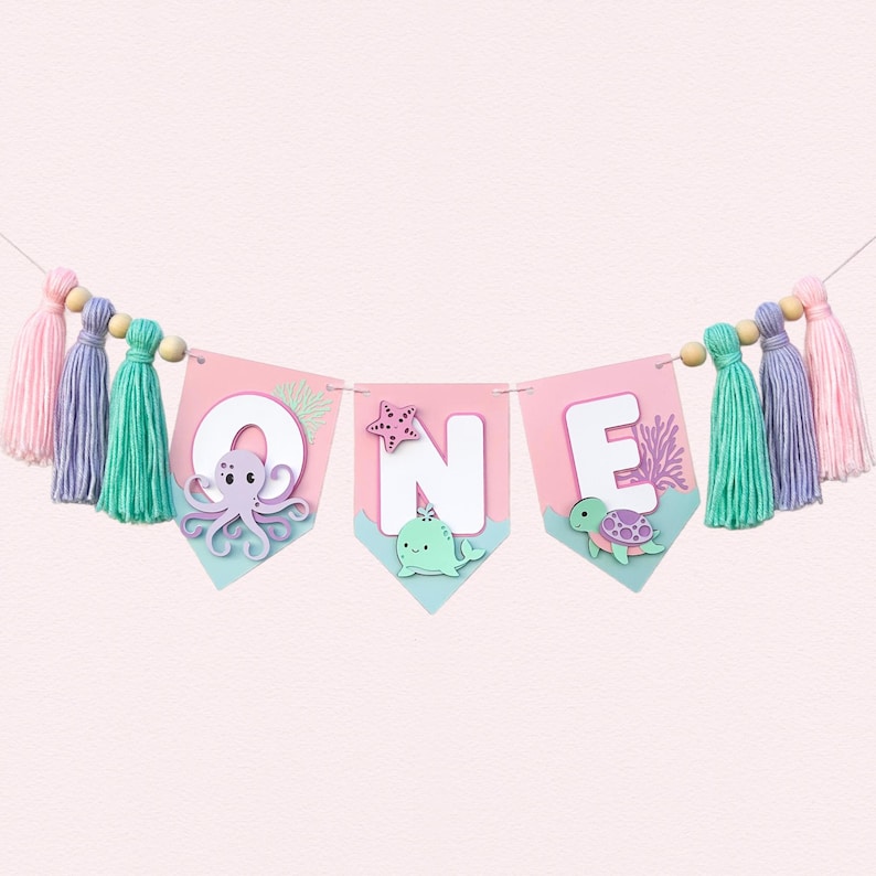 Pu&ograve; includere: Una ghirlanda rosa e blu con la scritta "ONE". Ogni lettera &egrave; su un gagliardetto rosa con disegni di creature marine. La ghirlanda &egrave; decorata con nappe e perline di legno, perfetta per un primo compleanno.