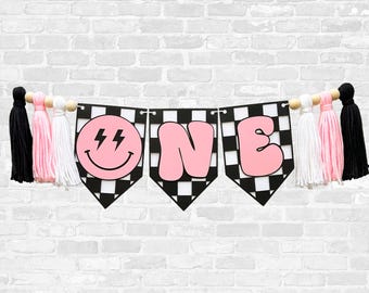 May include: A decorative banner featuring three pennants with a black and white checkered pattern. The pennants display a pink smiley face with lightning bolt eyes and the letters O, N, and E in pink. The banner is accented with black, pink, and white tassels.