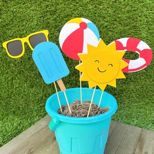 May include: A turquoise bucket filled with sand holds beach-themed cutouts. A yellow pair of sunglasses, a blue popsicle, a beach ball, a smiling sun, and a red and white life preserver are attached to wooden sticks.