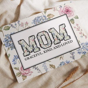 May include: A floral-themed greeting card with the word "MOM" in a bold, decorative font. The letters are filled with a floral pattern, and the card includes the text "GRACEFUL, KIND, AND LOVED". The background features a floral design with pink roses and blue hydrangeas.