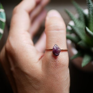 May include: A gold ring with a purple gemstone set in the center. The ring is worn on a person's finger.