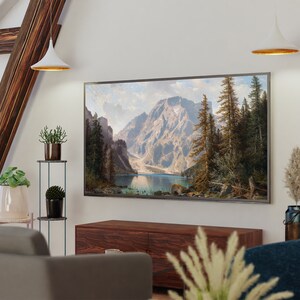 May include: A living room interior with a large framed landscape painting of a mountain lake and forest hanging above a wooden entertainment center. The painting is in a silver frame and is centered on the wall. There are two white pendant lights hanging from the ceiling above the painting. There are plants on a metal stand to the left of the painting.