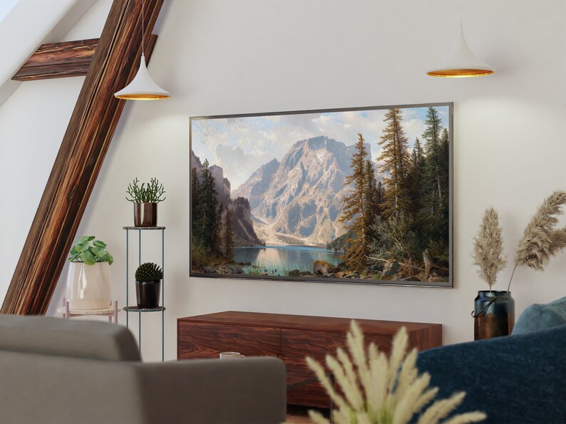May include: A living room interior with a large framed landscape painting of a mountain lake and forest hanging above a wooden entertainment center. The painting is in a silver frame and is centered on the wall. There are two white pendant lights hanging from the ceiling above the painting. There are plants on a metal stand to the left of the painting.