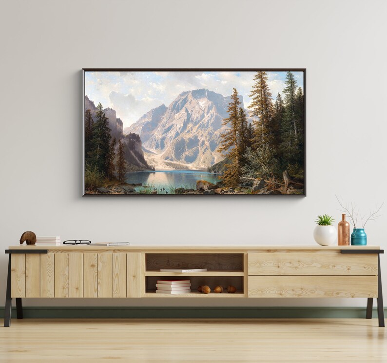 May include: A framed landscape painting depicting a mountain lake surrounded by trees and mountains. The lake is a light blue color and the mountains are a dark green color. The trees are a dark green color and the sky is a light blue color.
