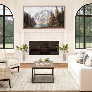 May include: A white living room with a white couch and two chairs. The room has a fireplace with a black screen and a large framed picture of a mountain scene above it. There is a coffee table in front of the couch with a few plants on it. The room has two large arched windows with a view of green trees and grass.