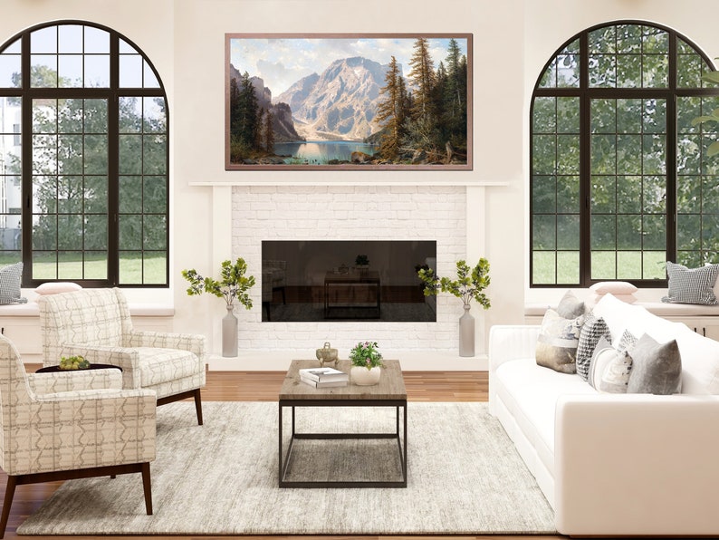 May include: A white living room with a white couch and two chairs. The room has a fireplace with a black screen and a large framed picture of a mountain scene above it. There is a coffee table in front of the couch with a few plants on it. The room has two large arched windows with a view of green trees and grass.