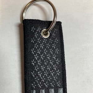 May include: A black and silver key chain with a silver ring and a black fabric strap with a gray embroidered American flag design.