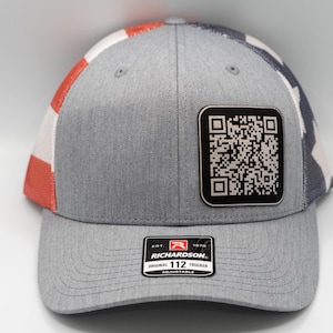 May include: A gray and red baseball cap with a white and blue American flag design on the side. The cap has a black square patch with a QR code on it. The cap also has a black label with the text "Richardson. 112 Adjustable" on the front.