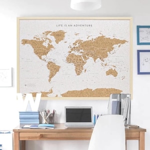 May include: A corkboard world map with the text "Life is an adventure" at the top. The map is hanging on a white wall above a wooden desk with a laptop, a white chair, and a stack of books.