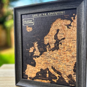 May include: A corkboard map of Europe in a black frame. The map is labelled with country names and has a title that reads "Life is an Adventure".