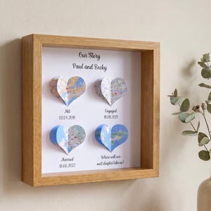 May include: A wooden framed artwork with four heart-shaped map cutouts. The text reads "Our Story", names, and dates of significant events. The frame is light oak. The artwork is a sentimental keepsake.