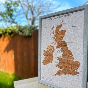 May include: A corkboard map of the United Kingdom with the text "Life is an Adventure" at the top. The map is framed in a light grey frame and is sitting on a light wood surface.