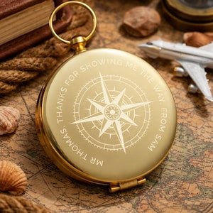 May include: A round gold-coloured metal compass with a circular ring at the top. The lid features an engraved compass rose design and the text: MR THOMAS, THANKS FOR SHOWING ME THE WAY, FROM SAM. The surface is smooth and reflective.
