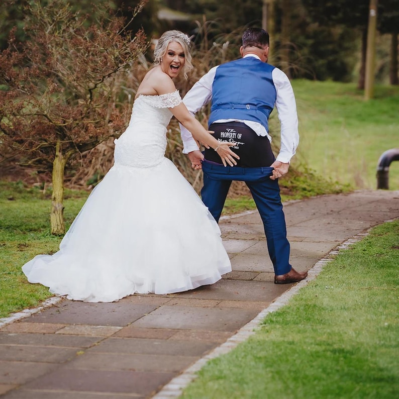 May include: A white off-the-shoulder wedding dress with a lace bodice and a full tulle skirt, paired with a blue vest and blue trousers. The trousers are pulled down to reveal black underwear with the white text Property of Mrs. Dapper.