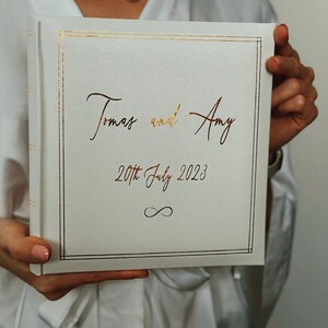 May include: A square white photo album with a gold foil border and gold foil text that reads Tomas and Amy, 20th July 2023, and an infinity symbol. The cover has a smooth, matte finish and a clean, minimalist design with elegant script lettering.