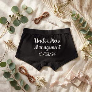 May include: Black boxer briefs featuring the white text Under New Management and the date 15/09/24 printed on the front. The garment has a wide elastic waistband and is displayed with decorative eucalyptus sprigs and a small gift box.