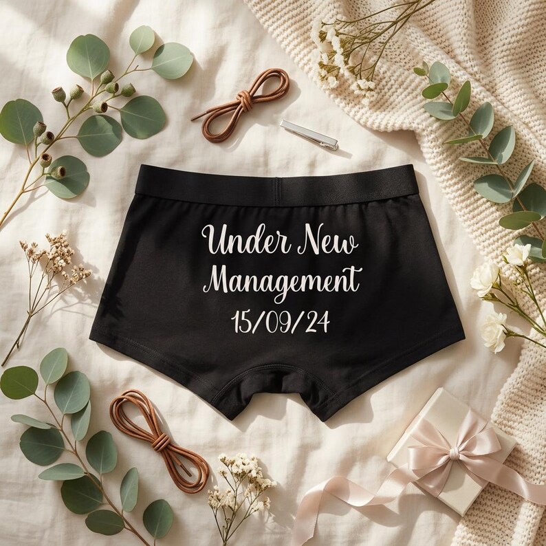 May include: Black boxer briefs featuring the white text Under New Management and the date 15/09/24 printed on the front. The garment has a wide elastic waistband and is displayed with decorative eucalyptus sprigs and a small gift box.