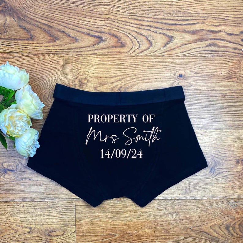 May include: Black boxer briefs featuring white printed text that reads PROPERTY OF Mrs Smith and the date 14/09/24. The garment has a wide elastic waistband and is displayed alongside a cluster of white peony flowers.
