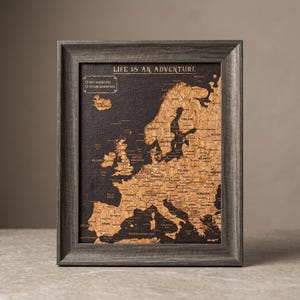 May include: A framed map of Europe featuring a cork texture for the landmasses against a dark background. The text LIFE IS AN ADVENTURE is printed at the top, with a legend for past and future adventures. The frame is a dark grey wood grain.