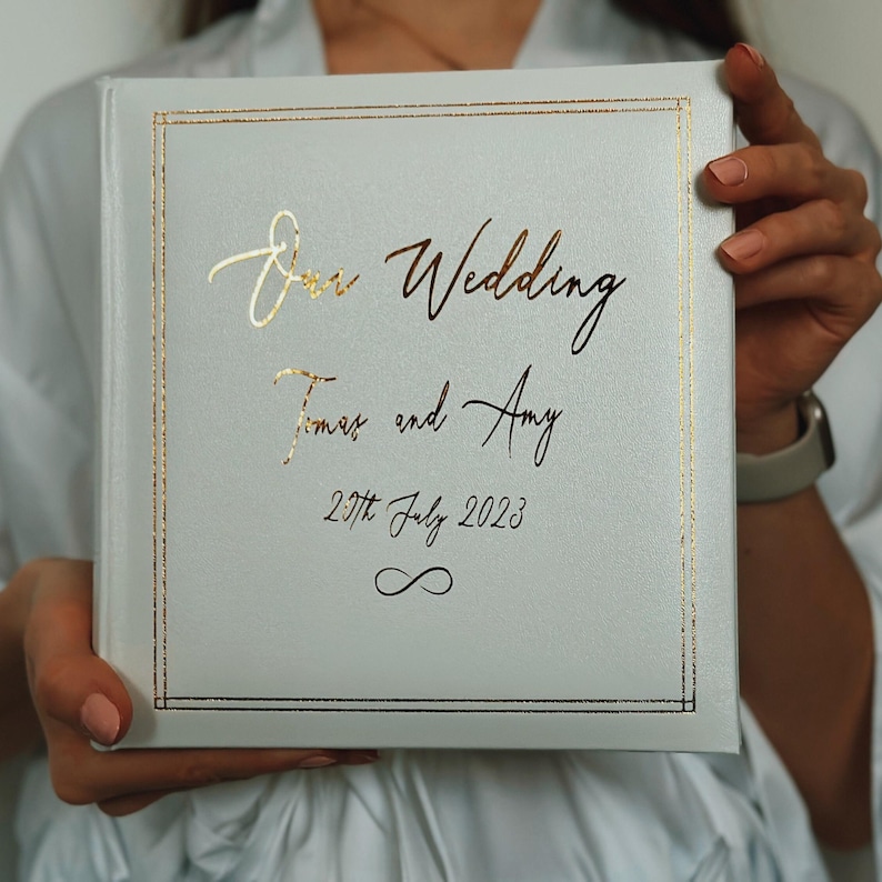 May include: A square white photo album with a gold foil border and gold lettering that reads Our Wedding, James and Amy, 29th July 2023, and an infinity symbol. The cover has a smooth, matte finish and a clean, minimalist design structure.