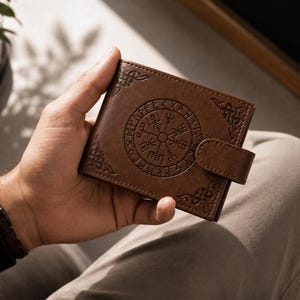 May include: A brown bi-fold wallet featuring an embossed circular Vegvisir symbol surrounded by a ring of runic characters. The wallet includes decorative corner patterns and a snap-button closure tab on the right side, with visible stitching.