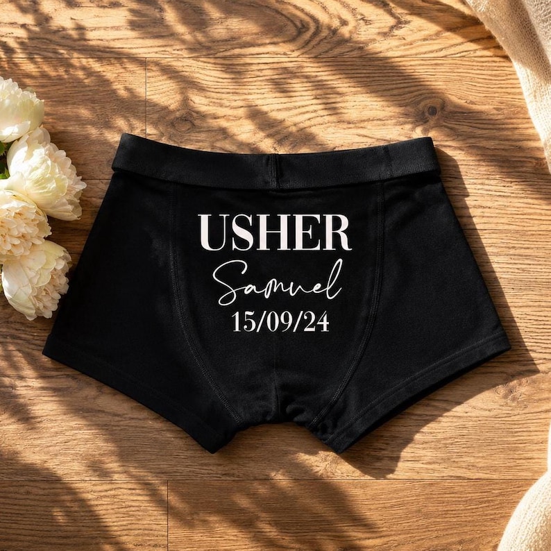 May include: Black boxer briefs featuring white printed text that reads USHER, followed by the name Samuel in a cursive script, and the date 15/09/24 below. The garment has a wide elastic waistband and visible stitching along the front panel.