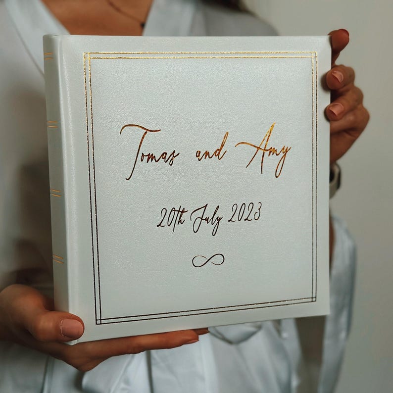 May include: A square white photo album with a gold foil border and gold lettering that reads Tomas and Amy, 20th July 2023, and an infinity symbol. The album has a smooth, matte finish and a spine with gold horizontal accent lines.
